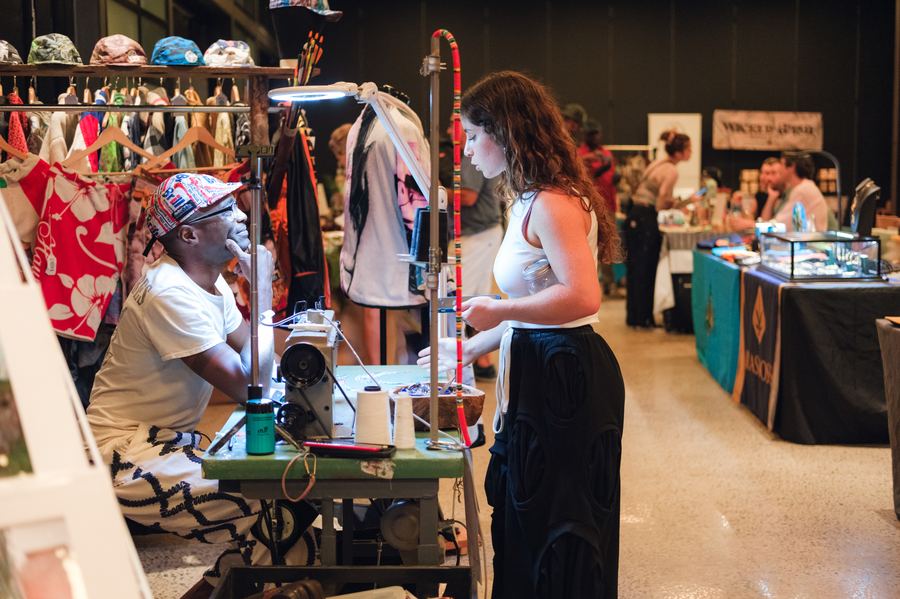 A visitor watching a maker work at a sewing machine among vintage clothing rails at a Mindful Matter Market